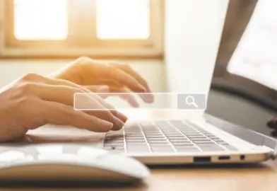 Close-up of hands typing on a laptop keyboard with a search bar overlay, illuminated by natural light from a nearby window.