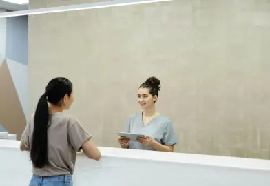 A healthcare professional at a reception desk smiling and holding a tablet while interacting with a patient standing in front of the desk.