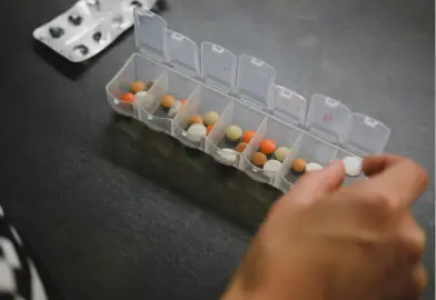 A hand reaching for a pill from a weekly medication organizer filled with various colored pills, with a blister pack of pills in the background.