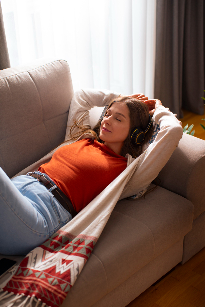 Patient relaxing on beige couch with hands behind head, wearing headphones and orange shirt, eyes closed peacefully during at-home ketamine therapy session