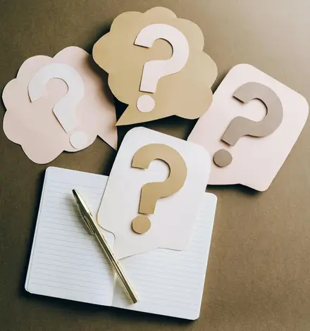 A pen and a notebook on a table, with paper question mark symbol cut outs scattered around the table