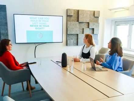 Three women sitting at a office table reviewing a presentation on a screen on the wall.