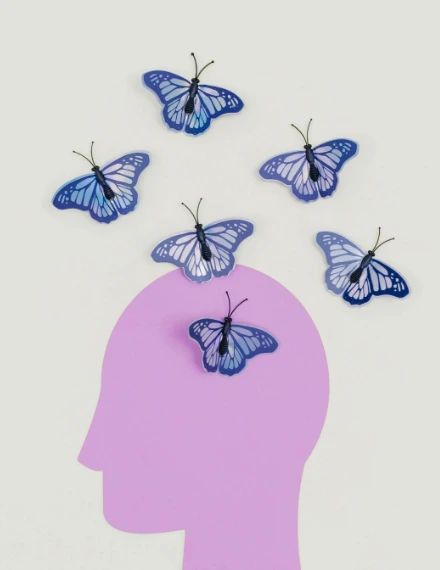 Side profile silhouette of a person's head in pink/purple, with six blue butterflies floating around it - one butterfly positioned inside the head silhouette and five others scattered in the surrounding beige background space.