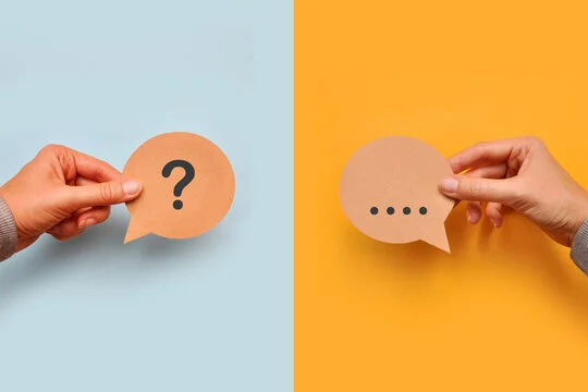 Two hands holding paper speech bubbles - one with a question mark on blue background, one with ellipsis dots on orange background