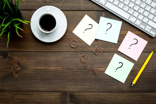 A wooden table with a cup of coffee, keyboard, and pencil surrounded by question mark symbols written on sticky notes.