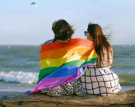 Two women sitting at the beach with a LGBTQIA+ flag wrapped around their shoulders.
