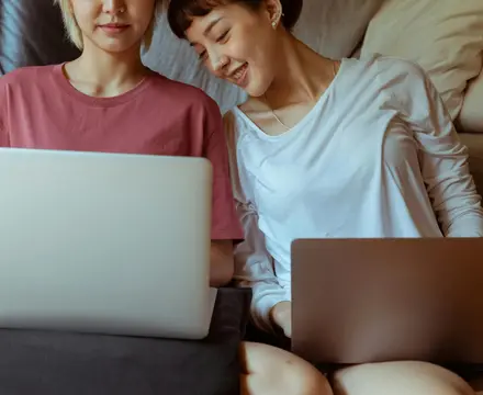 Two women sitting on a couch both with their laptops open looking for their therapist options.
