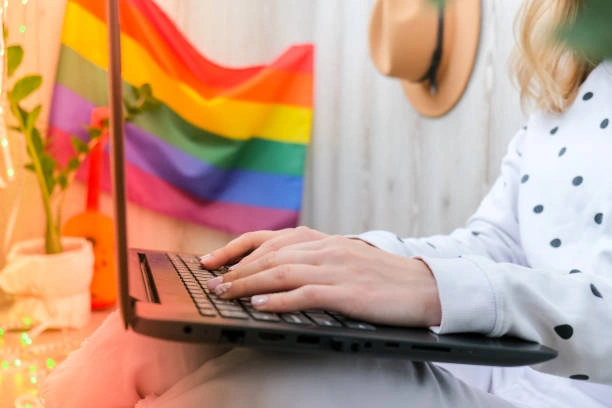 A person sitting on their laptop looking for their therapist options with a LGBTQIA+ flag in the background.