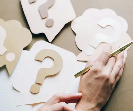 A pen and notebook sitting on a table with paper question mark cut outs scattered around the table.