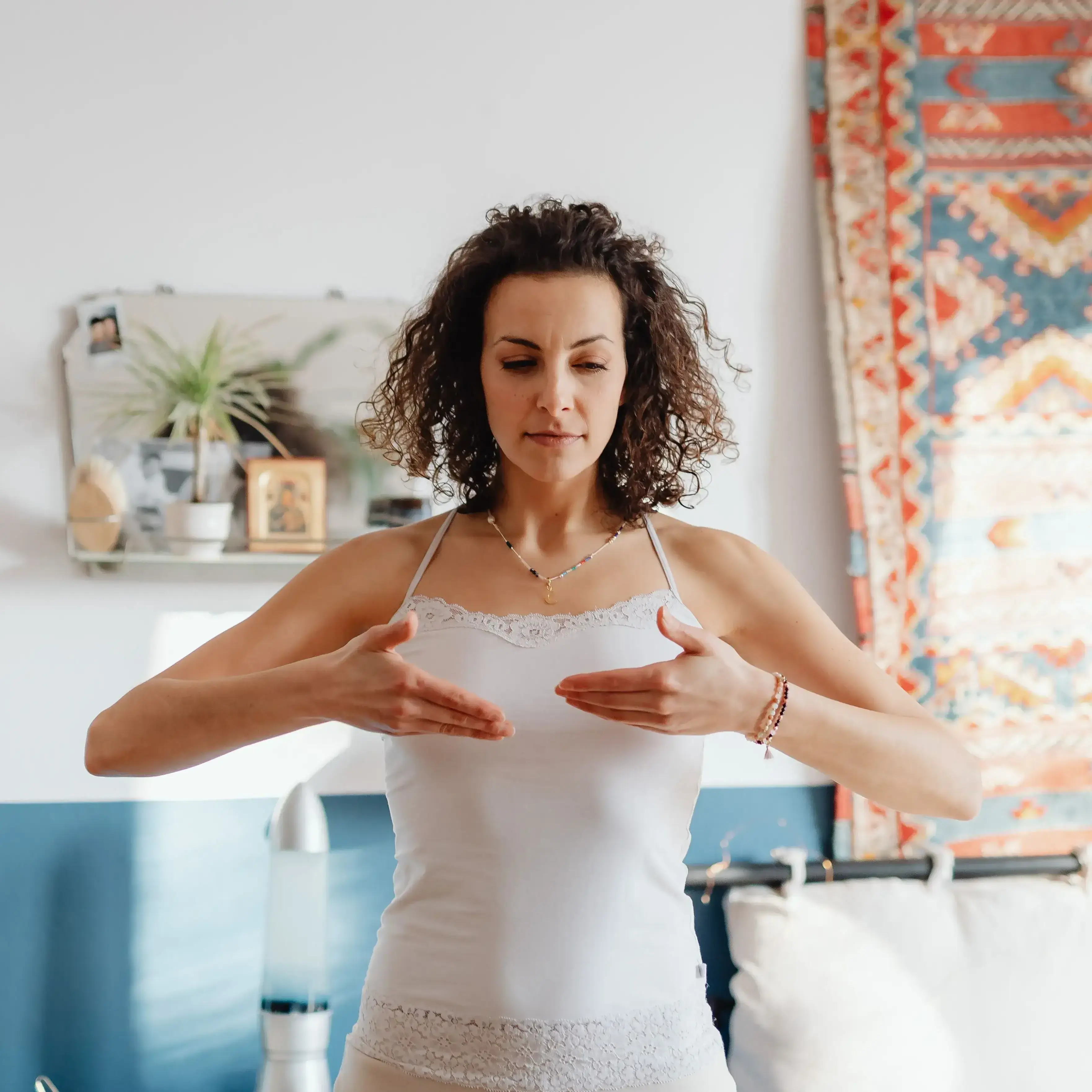 A woman standing in her bedroom with her eyes lowered and her hands held over her chest, practicing a breathwork exercise.