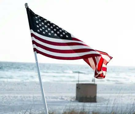USA flag waving in the wind with a ocean view in the background.