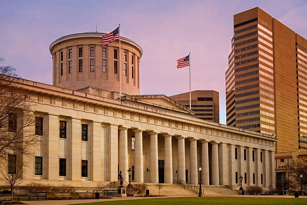 Ohio state capitol building with a USA flags waving in the wind and a sunset in the background.