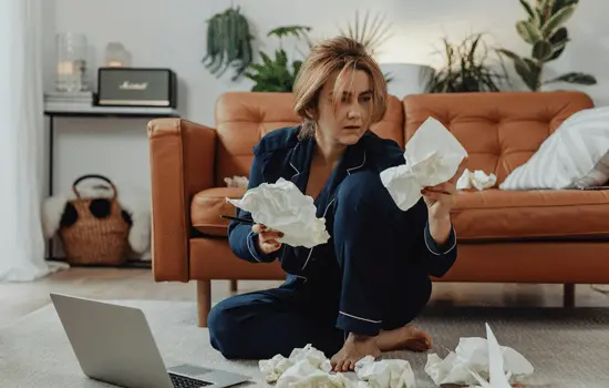 Woman sitting cross-legged on floor surrounded by crumpled papers with laptop open, appearing overwhelmed while working from home