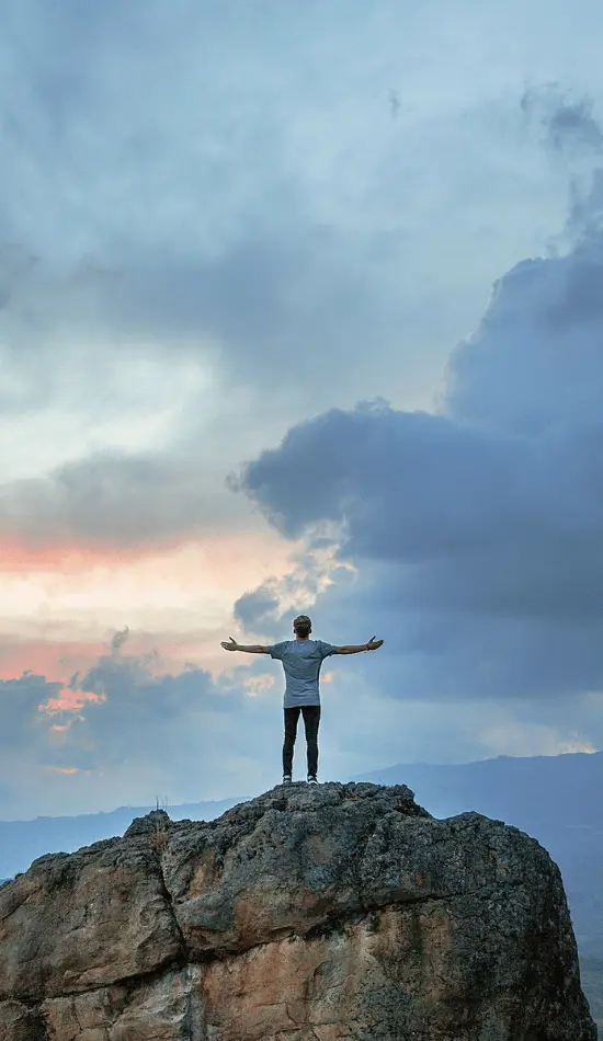 Person standing triumphantly with arms outstretched on rocky mountain peak against dramatic cloudy sky, symbolizing overcoming bipolar disorder challenges
