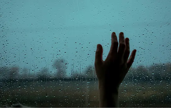 Silhouette of hand pressed against rain-covered window with water droplets, conveying feelings of isolation and sadness associated with depression