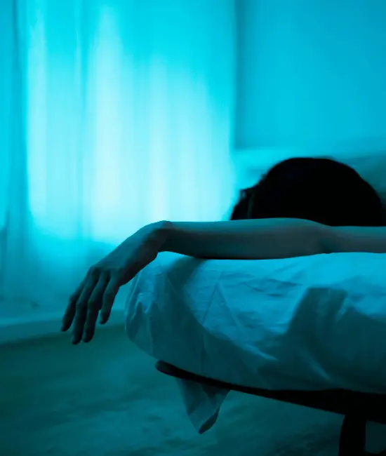 Person lying in bed in blue-tinted lighting with arm hanging over edge, illustrating fatigue and hopelessness common in depression