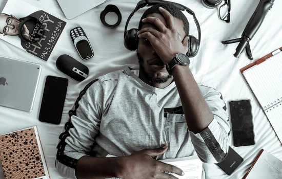 Overhead view of person lying on floor surrounded by scattered electronic devices, papers and technology, appearing overwhelmed by modern life demands