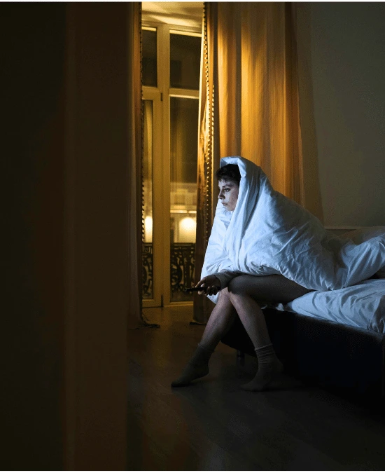 Person sitting on bed in dark bedroom illuminated by warm golden light from window, unable to sleep and experiencing insomnia