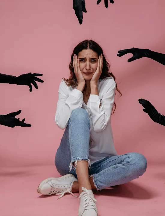 Woman sitting cross-legged holding her head in her hands against pink background with dark hand silhouettes reaching toward her, representing feelings of panic and being overwhelmed