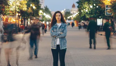 Woman standing alone and isolated in busy pedestrian area with blurred crowd moving around her, representing feelings of social disconnection and anxiety in public spaces