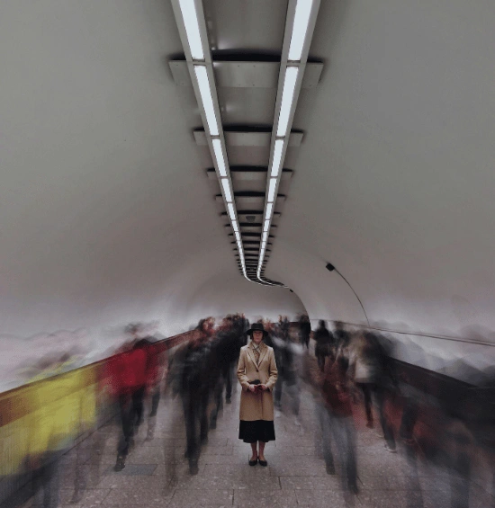 Lone figure standing in blurred subway tunnel with motion blur of passing people, representing feeling overwhelmed and isolated in crowded social spaces