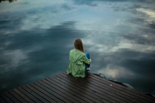 Person in green jacket sitting alone on wooden dock by calm water under cloudy sky, representing solitude, contemplation, and the withdrawn feelings associated with depression