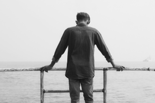 Black and white image of person standing alone at railing overlooking water, conveying persistent feelings of isolation and sadness despite previous treatment attempts